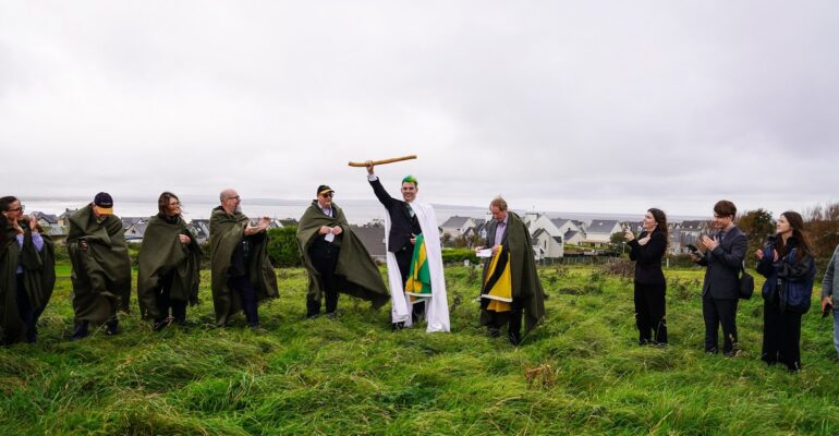 Sean O'Dowda Stephens raises the White Wand on the inauguration mound at Enniscrone, 9 October 2025, as cloaked clan members look on