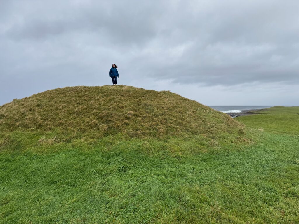 Carn Inghine Briain (Coggins' Hill) — a grass-covered earthen inauguration mound on Aughris headland, Co. Sligo, with the Atlantic Ocean visible to the right. A visitor stands on the tapering summit.