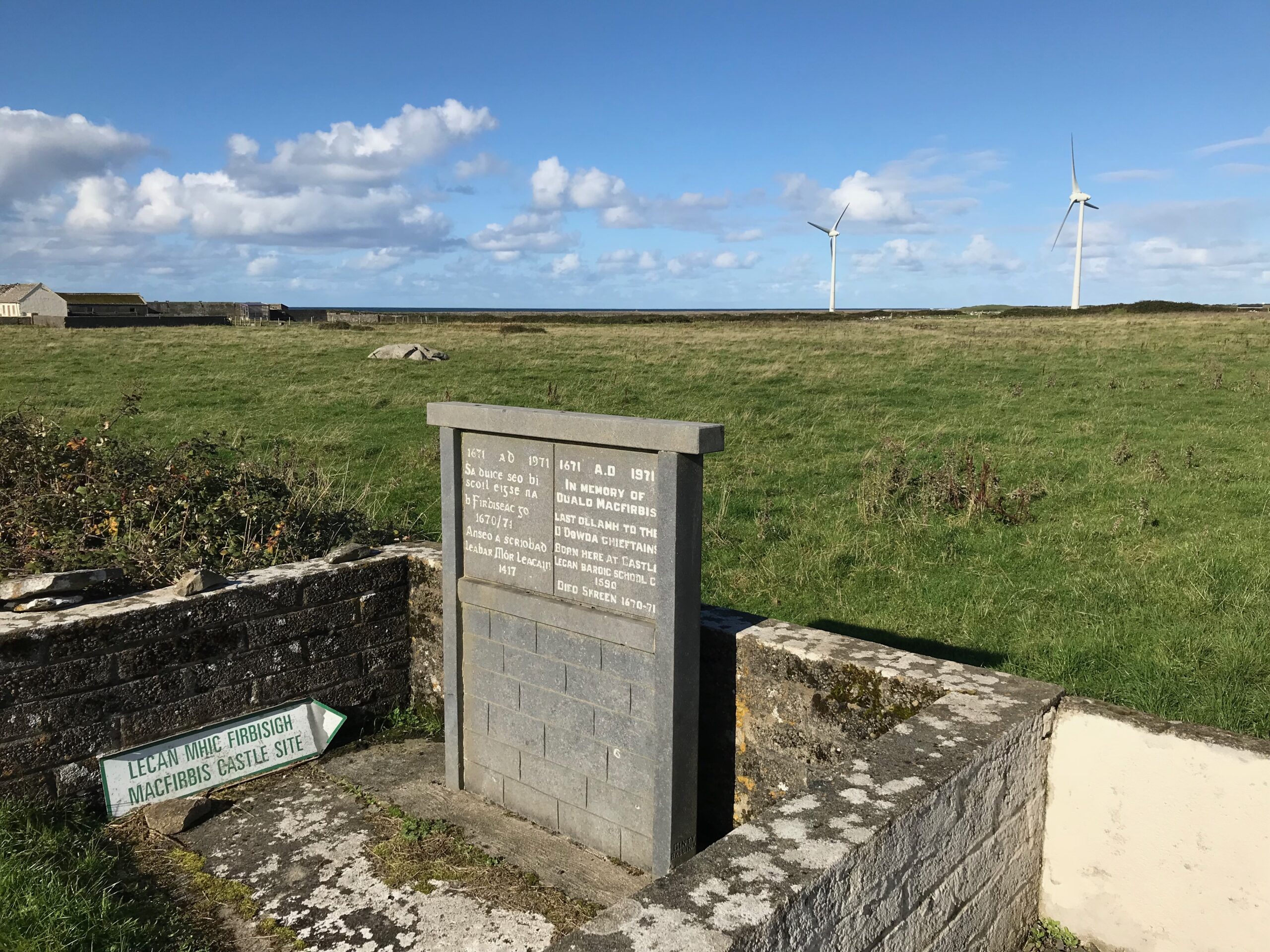 The memorial stone and road sign for Lecan Mhic Fhirbhisigh at Carrowhubbock North, Kilglass, with the Atlantic and wind turbines in the distance